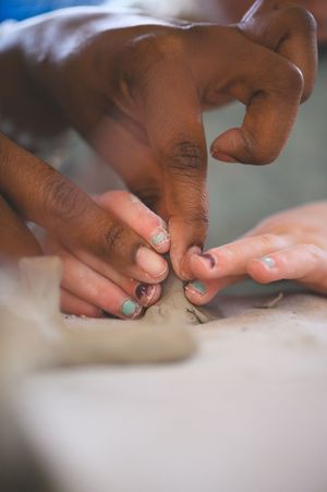 Hands working together shaping clay on a pottery wheel, with teal painted nails visible during a collaborative art class.