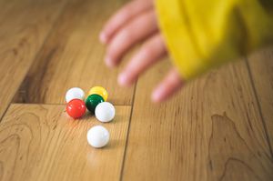 Child's hand reaching for colorful marbles on a wooden floor, including red, green, and white spheres.