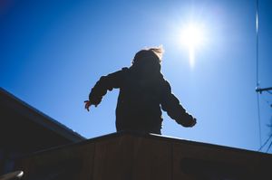 Silhouette of a child standing on a rooftop with arms outstretched against a bright blue sky and shining sun.
