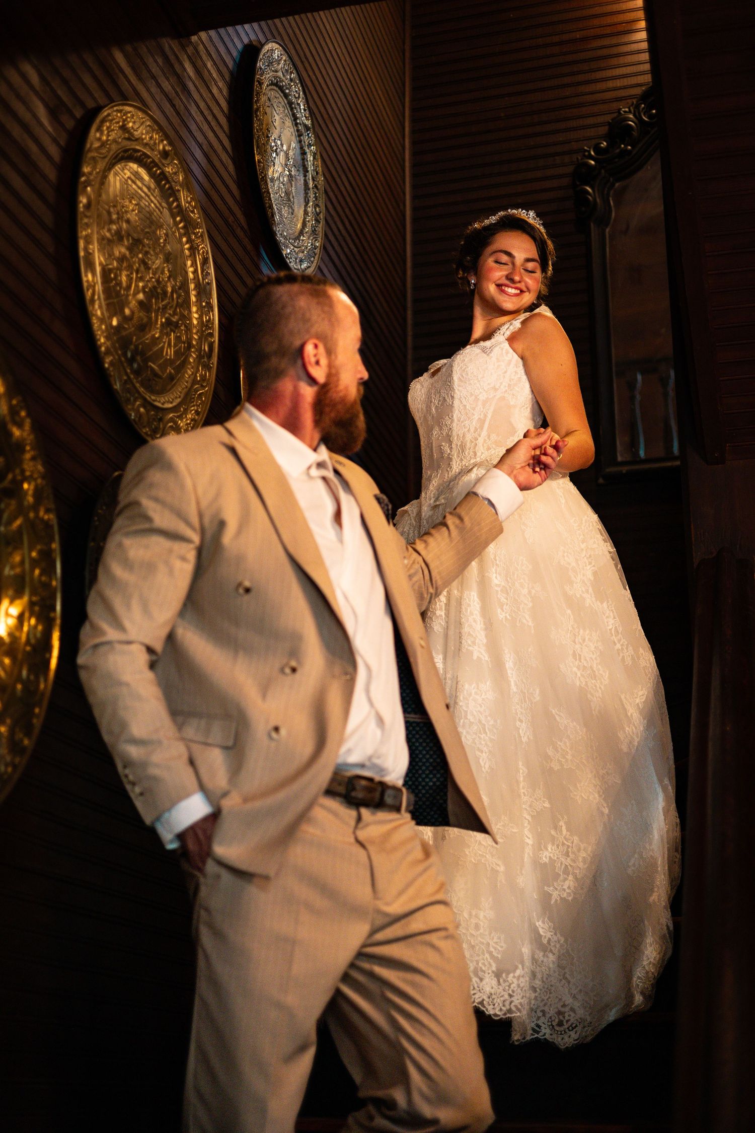 A groom in a tan suit holds his bride's hand as she stands elevated, smiling in a white lace wedding gown.