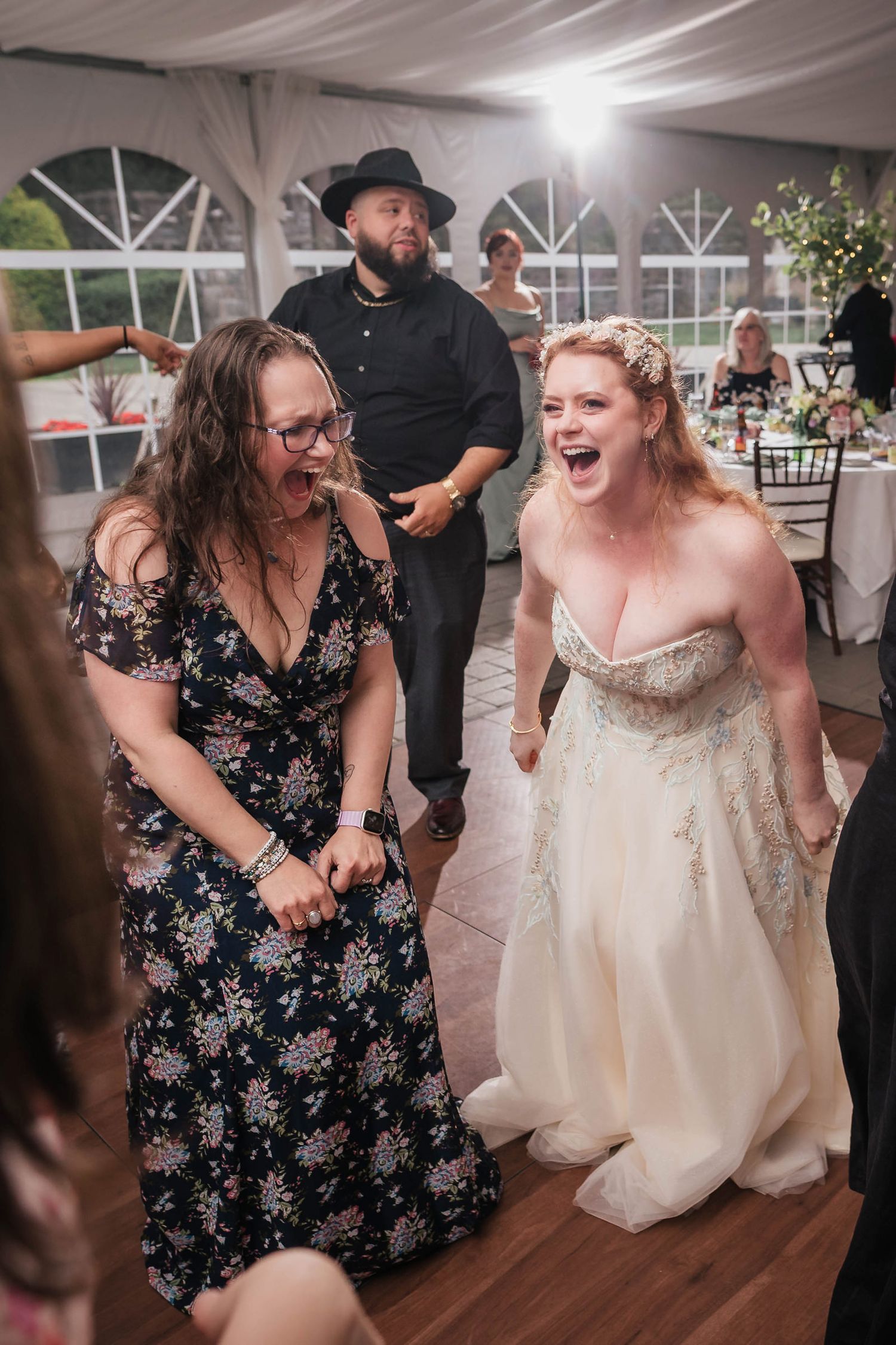 Bride laughing loudly with a guest on the dance floor, a man in a black hat behind them.