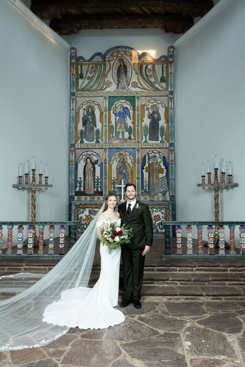 Bride and groom pose before ornate mosaic altarpiece in historic church, bride wearing cathedral veil and holding bouquet.