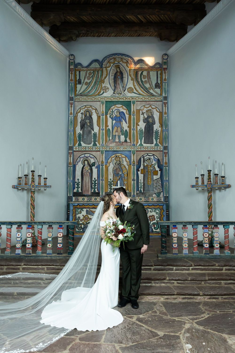 Bride and groom share a kiss before a colorful painted altar in a historic adobe church wedding ceremony.