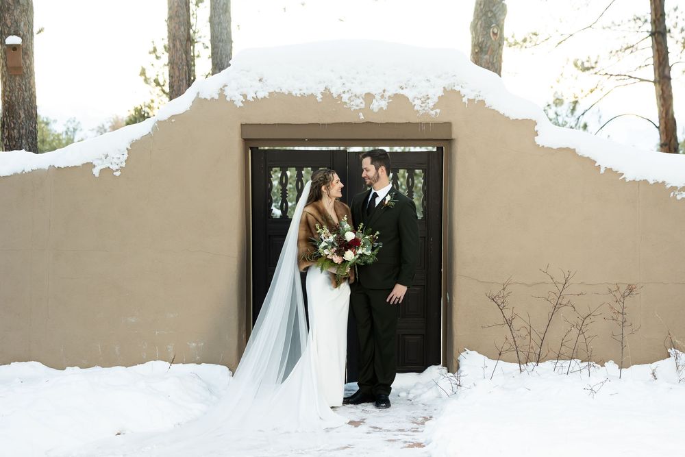 Bride and groom share a romantic winter moment in snowy setting outside rustic adobe building.