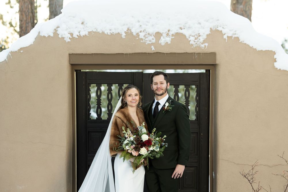 Bride and groom pose in winter wedding attire before snow-dusted adobe building, bride holding red bouquet with fur wrap.