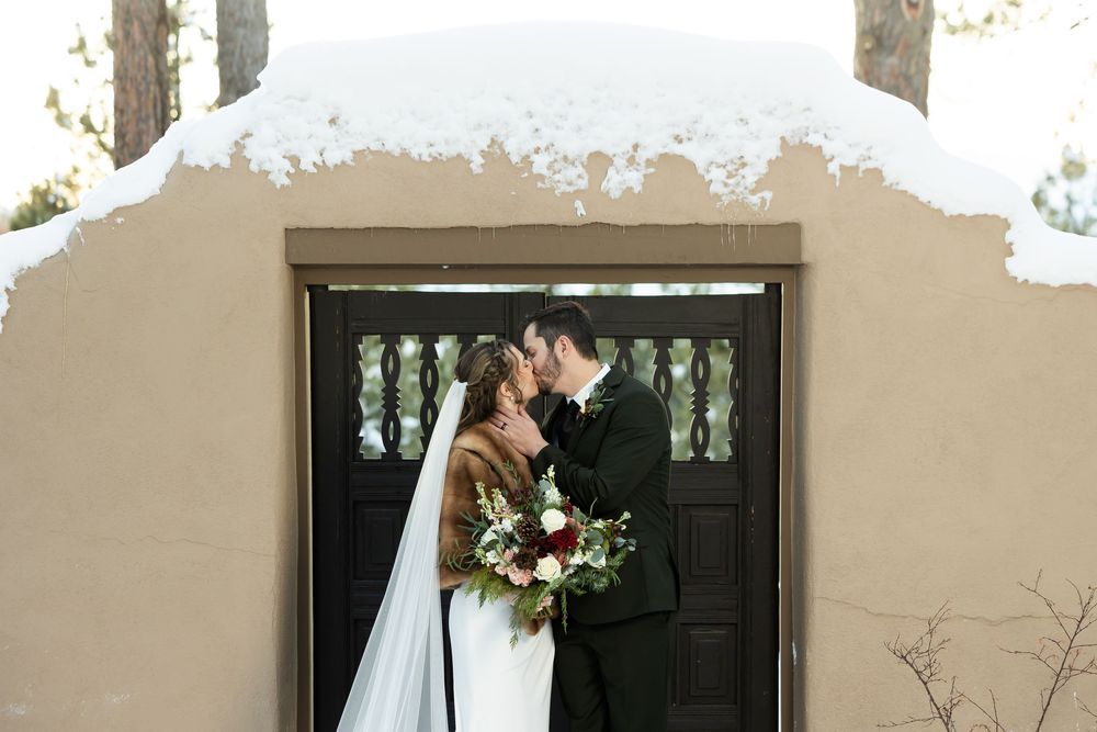 Bride and groom kissing in front of a snow-covered adobe chapel entrance in a winter wedding.