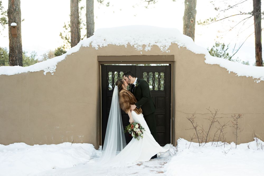 Bride and groom kissing in winter snow outside rustic adobe building with dark wood door and snow-covered roof.