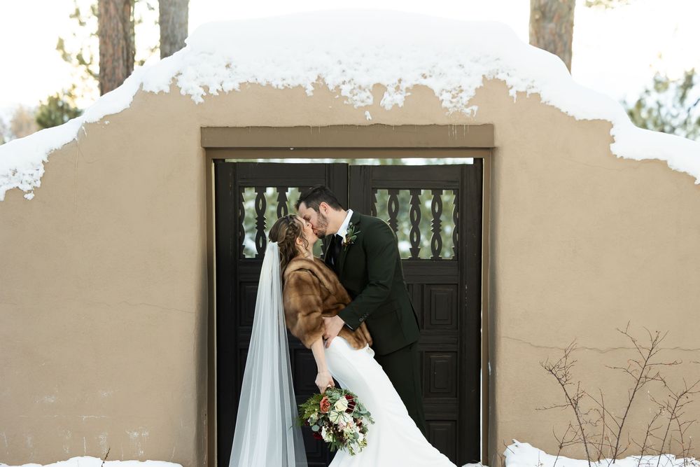 Bride and groom share a kiss in the snow outside a rustic adobe chapel, bride wearing a fur stole and veil.