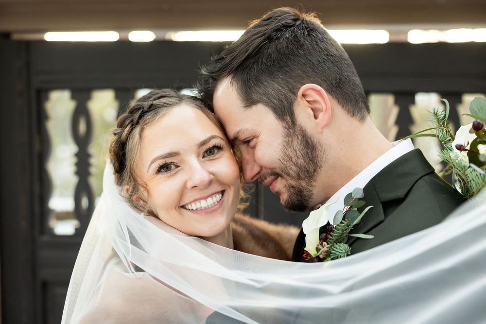 A smiling bride in a veil leans close to her groom in a tuxedo with a floral boutonniere on their wedding day.