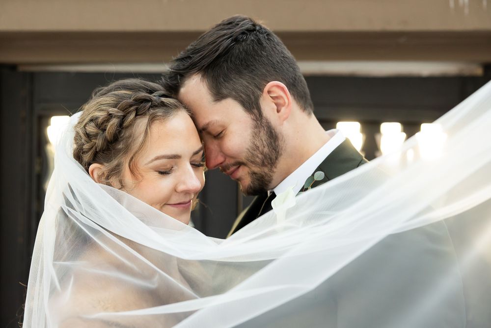 Bride and groom touch foreheads tenderly under a flowing veil, bride wearing braided updo on their wedding day.