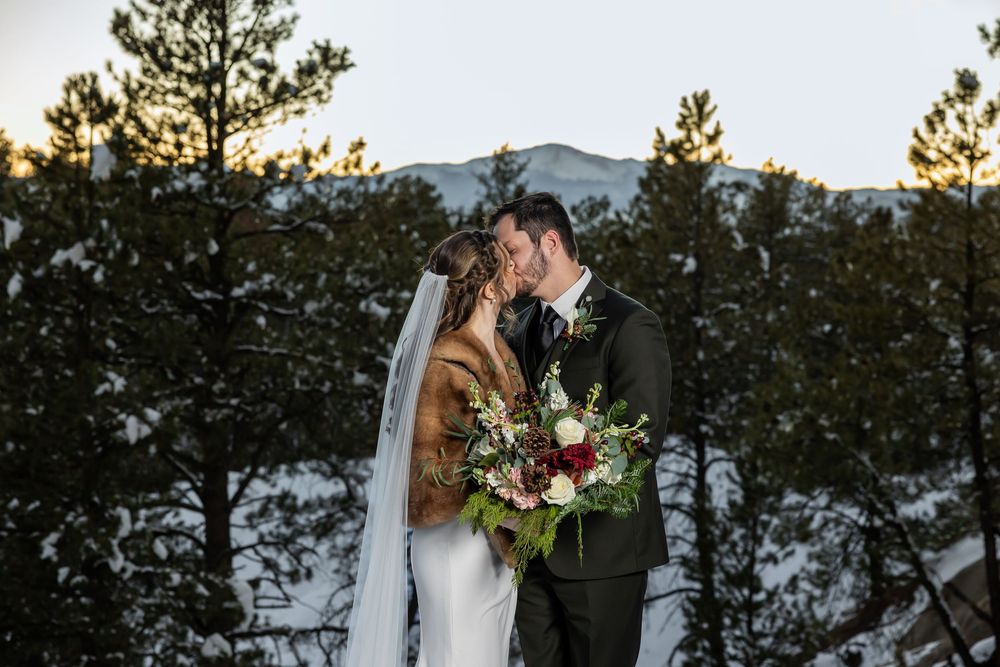 Bride and groom share a kiss in a snowy mountain forest, bride holding a red and green bouquet, at sunset.
