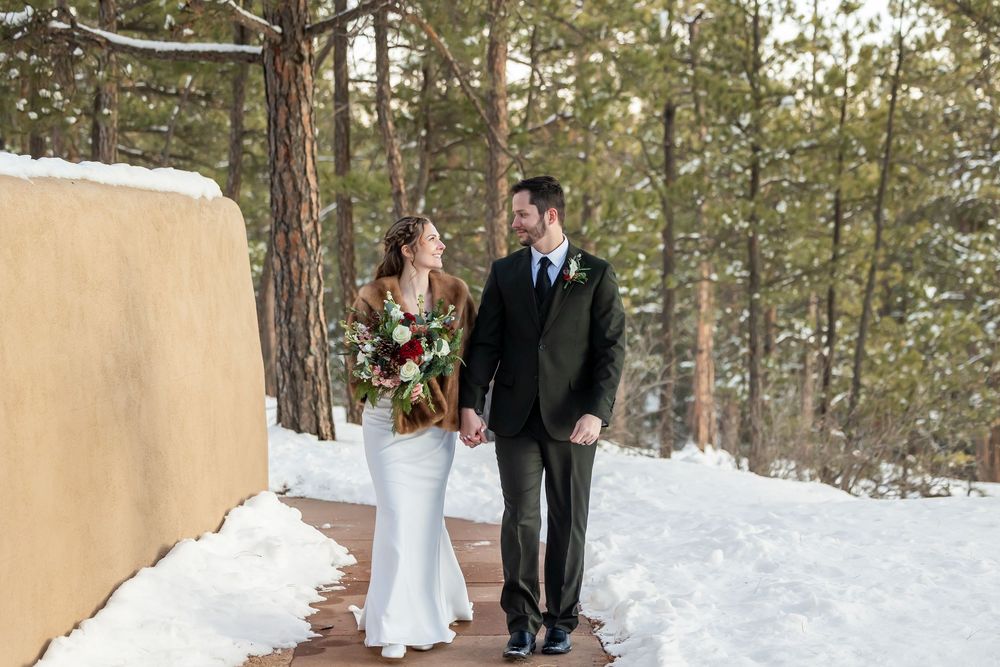 Bride and groom walk hand-in-hand on snowy winter path surrounded by pine trees, bride holding red bouquet.