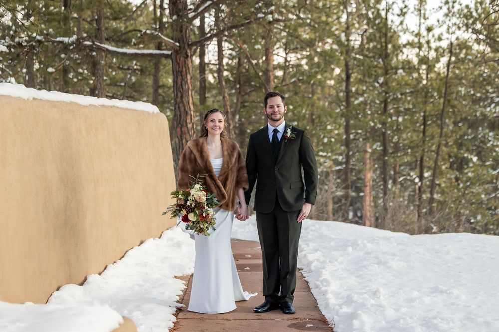 Couple in winter wedding attire holding hands on a snowy wooden path surrounded by pine trees and adobe wall.