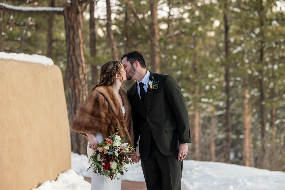 Bride and groom sharing a kiss in a snowy winter forest setting, bride wearing fur stole and holding bouquet.