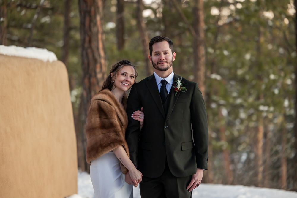 Bride in fur stole and groom in dark suit pose together outdoors in snowy winter forest wedding setting.