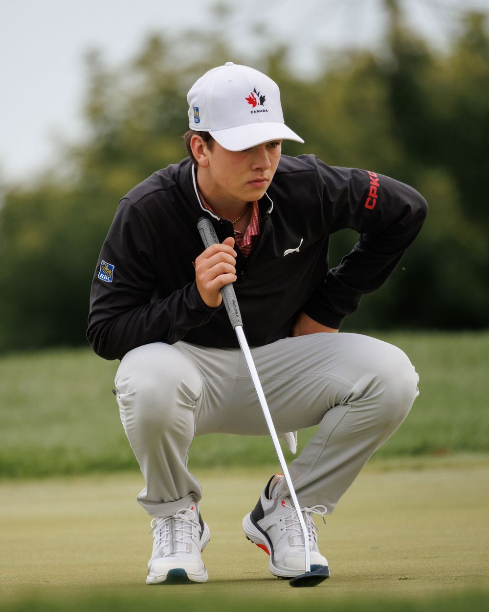 Junior golfer in Golf Canada hat crouches to read the green with putter.