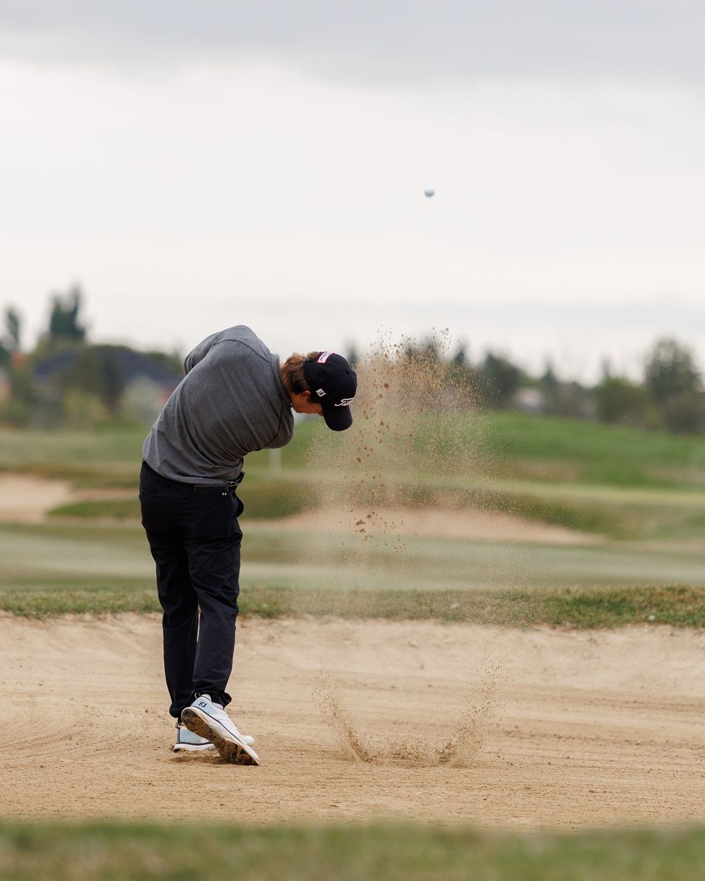 Golfer blasts out of a sand bunker sending sand flying on the course.