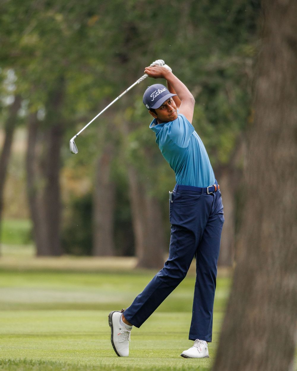 Golfer in teal shirt completing a full swing follow-through on a tree-lined fairway.