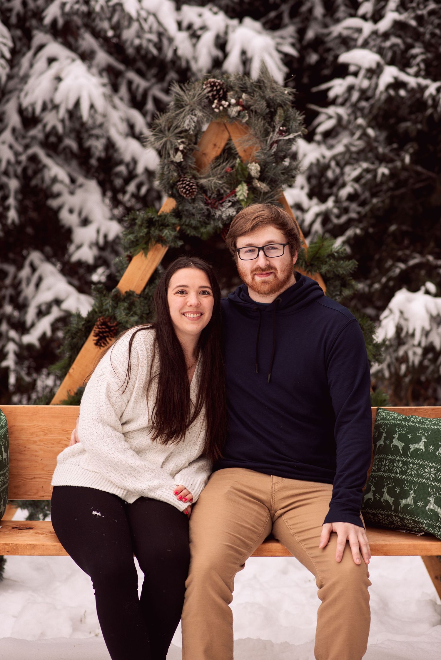 Couple smiling together on a wooden bench with a Christmas wreath, surrounded by snow-covered pine trees.