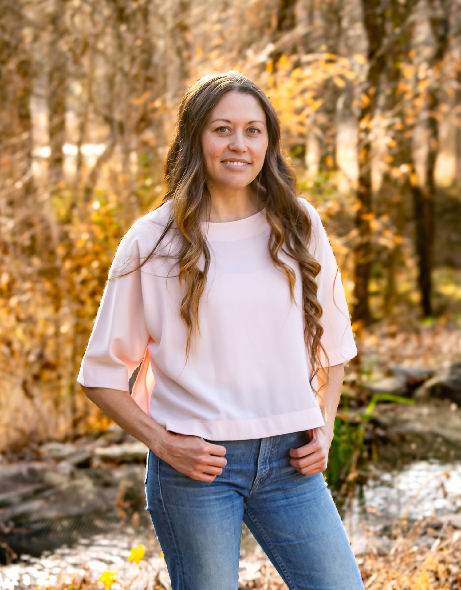 Woman in pink top and jeans smiling outdoors among autumn trees near a stream.