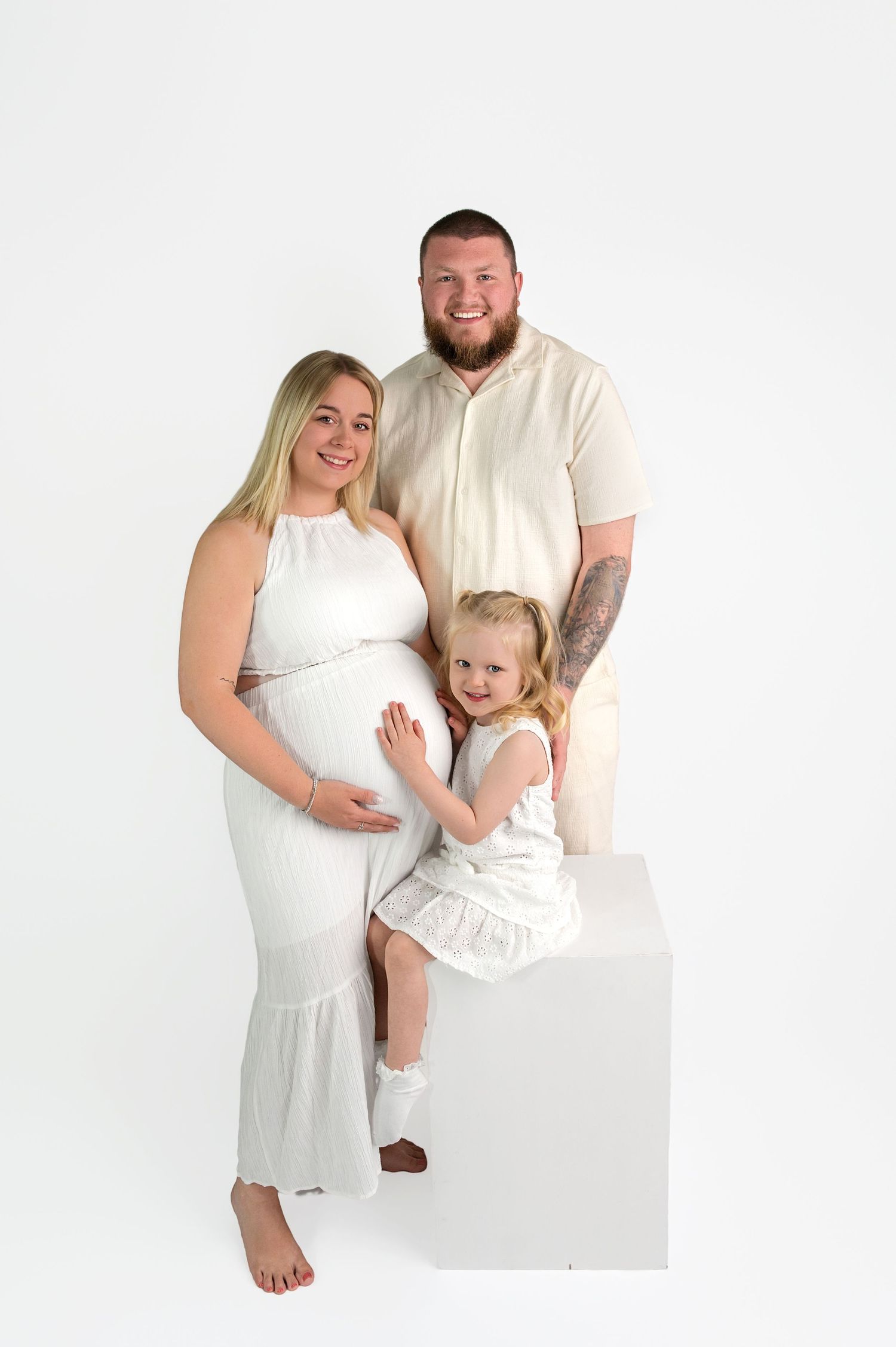 Pregnant woman in white dress poses with bearded husband and young blonde daughter in bright studio maternity photo.