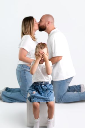 A young child covers her eyes as her parents share a kiss behind her in a bright white studio setting.