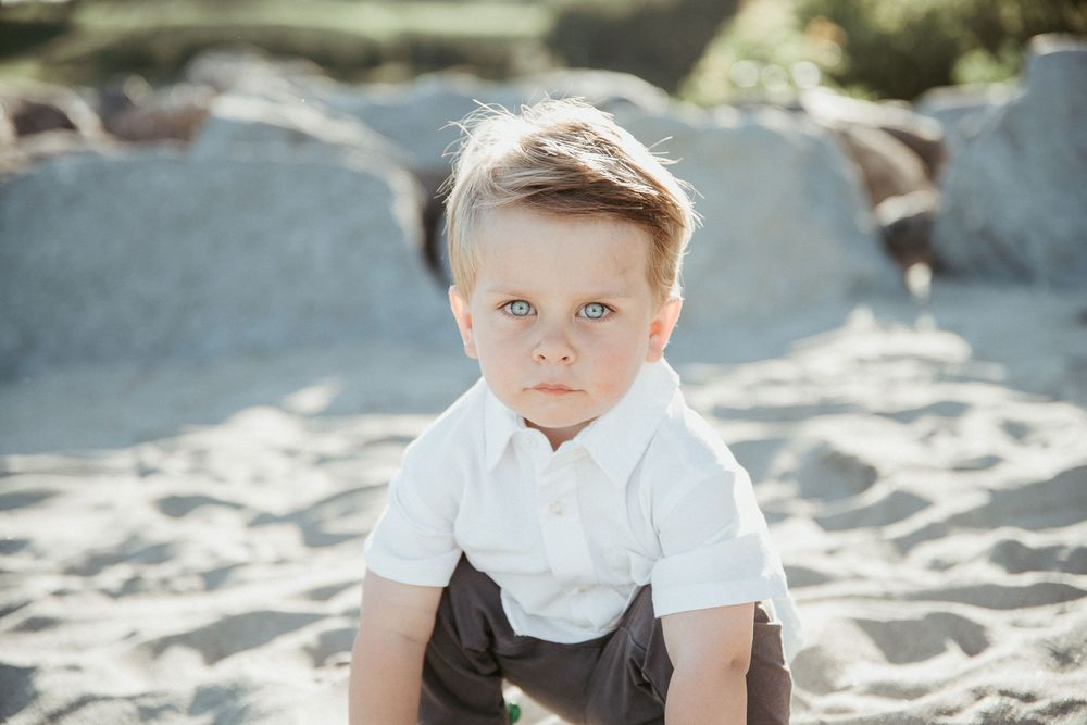 Young toddler boy with blue eyes in white shirt sitting on sandy beach, looking at camera.