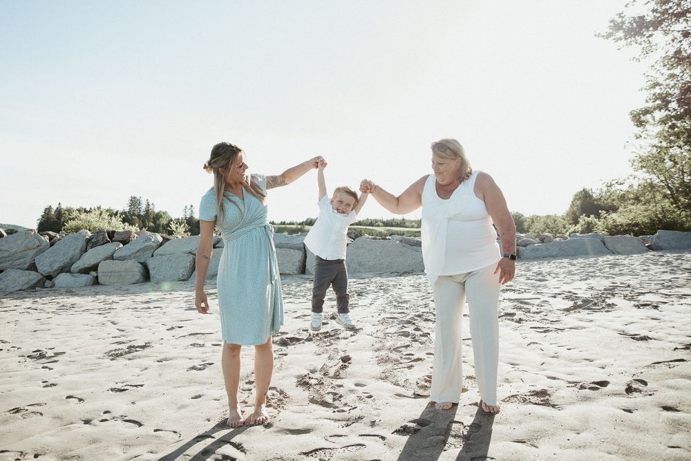 Two women and a child hold hands and play on a sunny sandy beach near large rocks and trees.