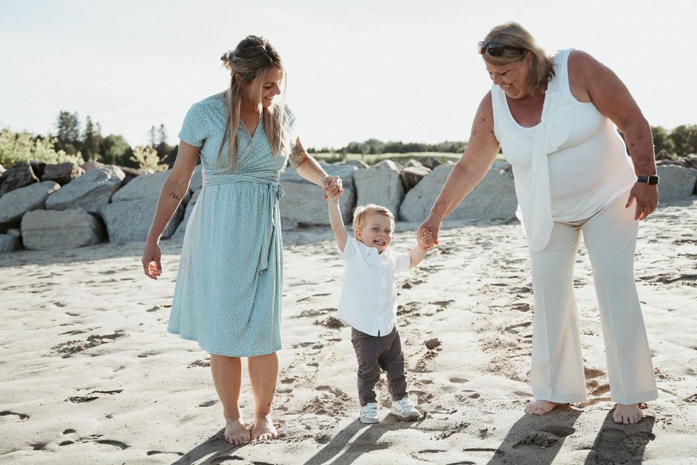 Two women help a toddler walk on a sandy beach, one in a light blue dress and the other in white pants.
