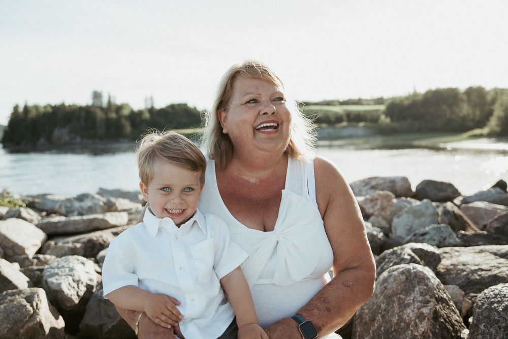 Grandmother and young boy smiling on rocky shoreline with water and trees in background on sunny day.
