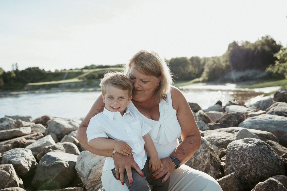 A grandmother and young boy sit together on rocky shore, sharing a warm embrace with a sunlit river in the background.