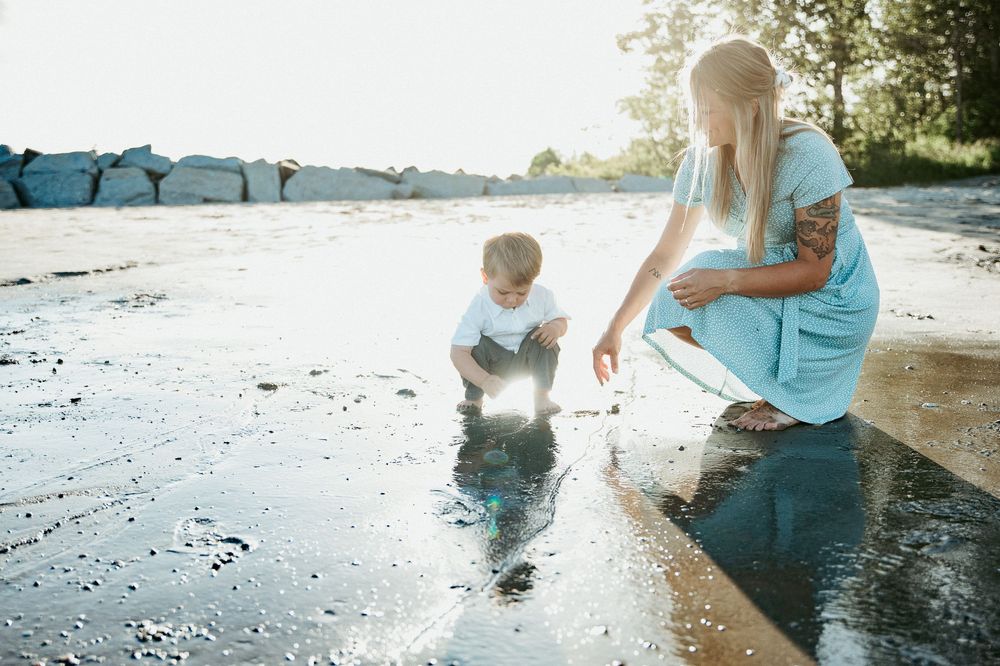 A woman and young boy crouch together near a shallow puddle outdoors, enjoying a sunny day by a rocky shoreline.