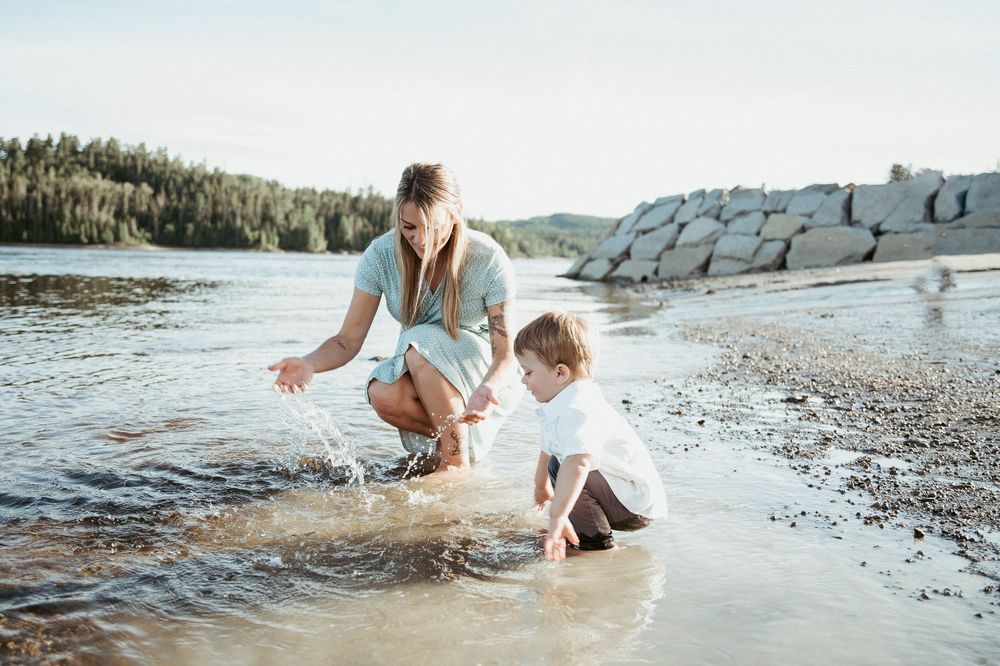 A mother and child playing in shallow water on a sunny beach with rocky shores and green trees.