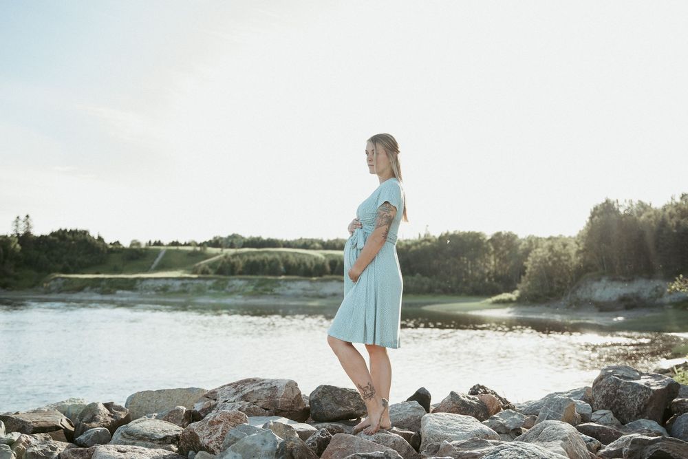 A young woman in a light blue dress standing on rocks by a serene river on a sunny day.