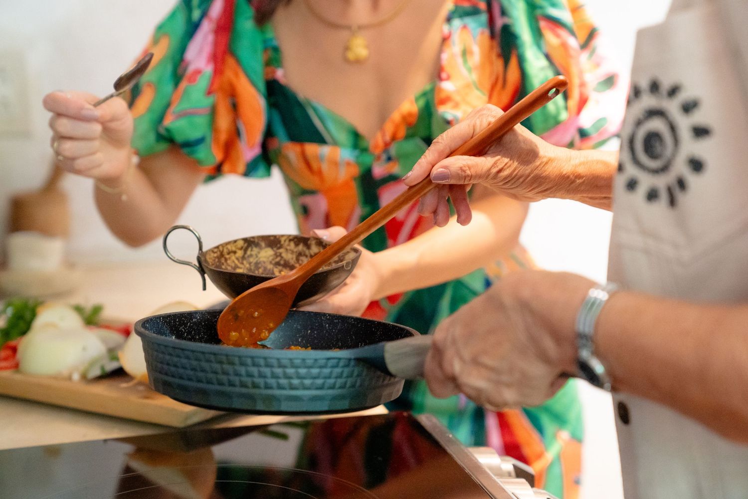 Two people cooking together, stirring a savory dish in a blue pan using wooden spoons in a bright kitchen.