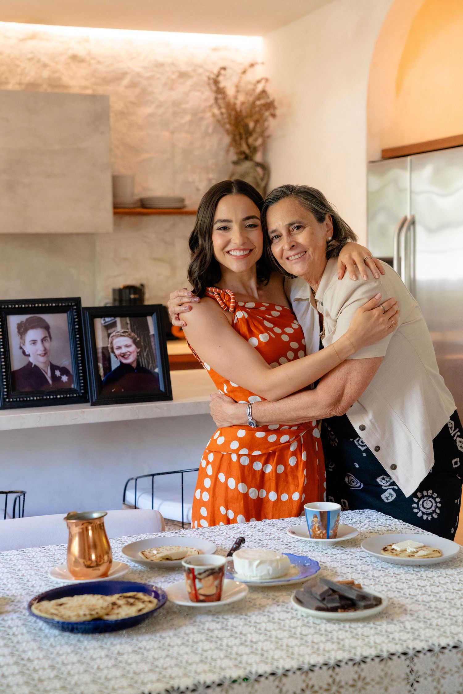 Two women embrace warmly at a kitchen table set with food and drinks, framed photos visible in the background.