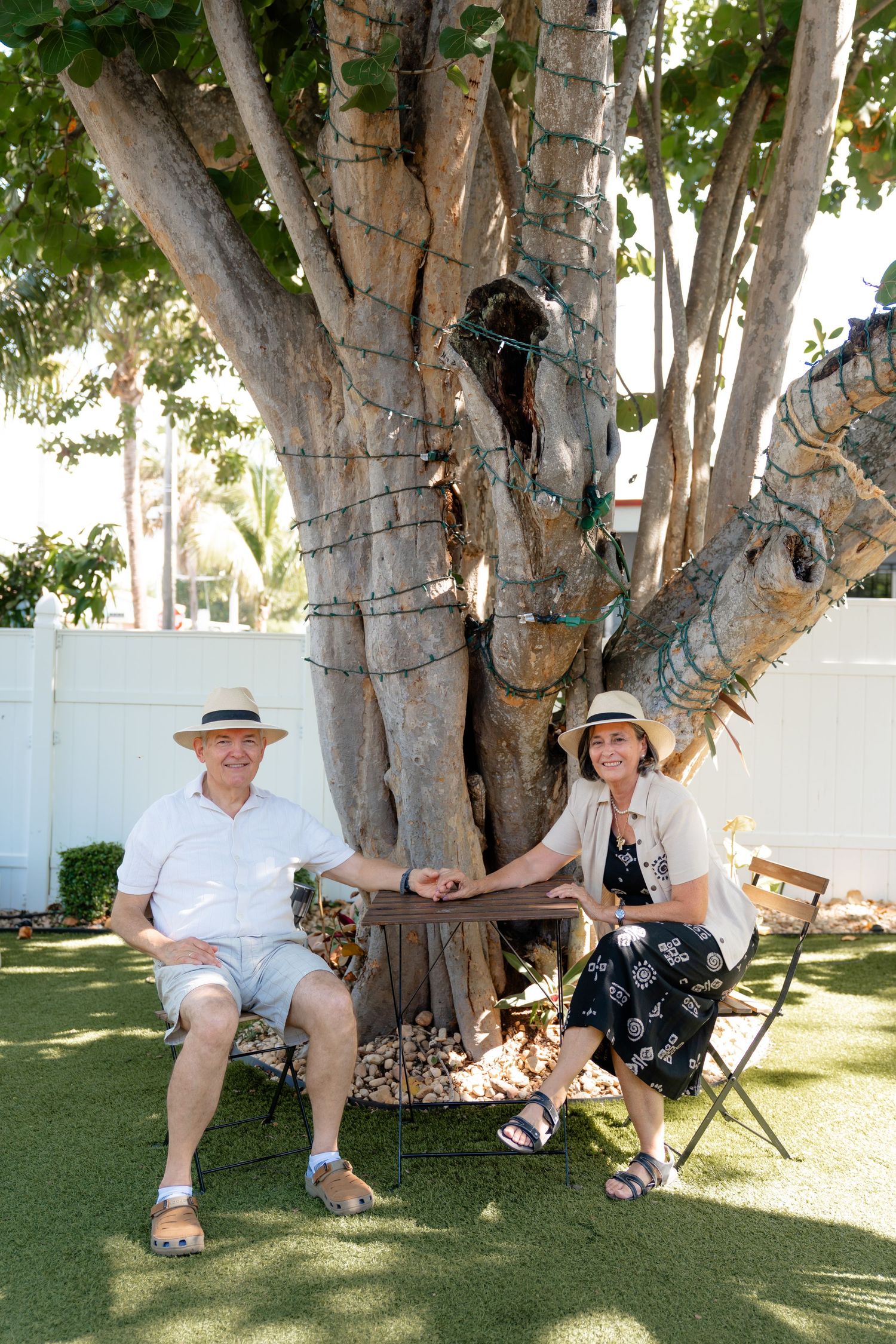 A couple in straw hats sitting on chairs beside a large tree in a sunny backyard garden.