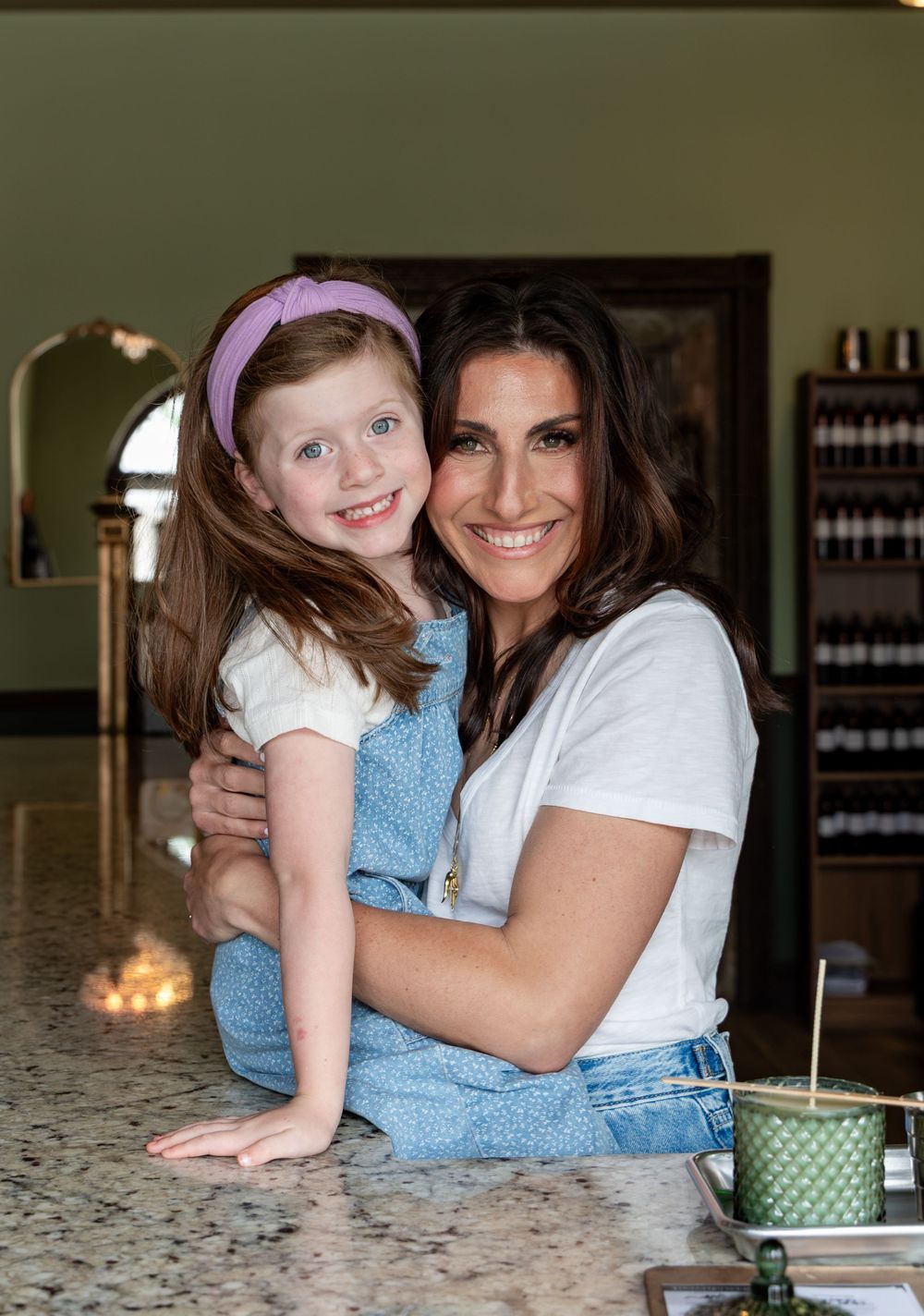 A woman and young red-haired girl in denim overalls smile together at a kitchen counter indoors.