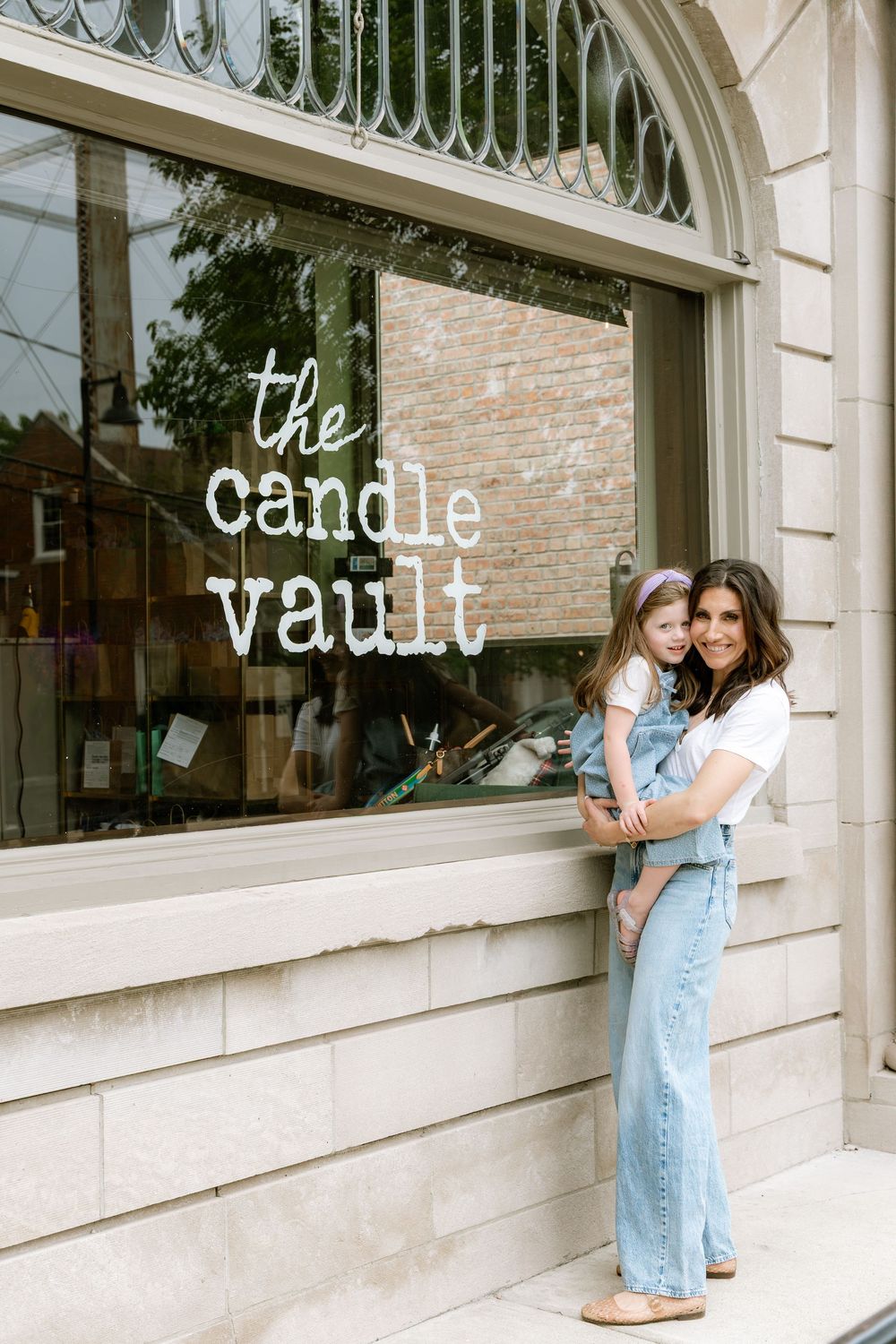 A woman holding a young girl smiles in front of The Candle Vault shop window in a charming downtown storefront.