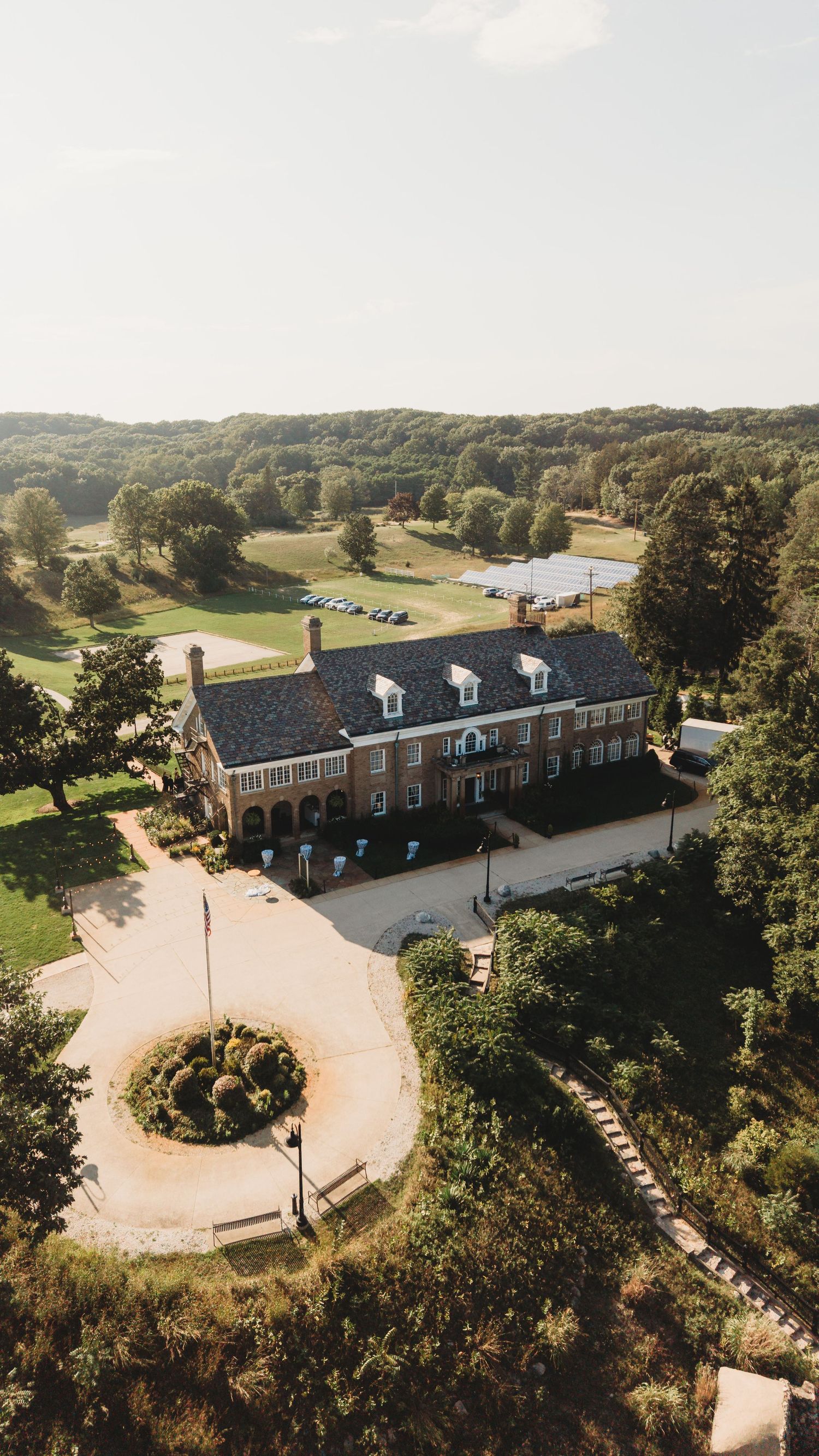 Aerial view of a grand brick estate surrounded by lush green trees and manicured grounds with a circular driveway.