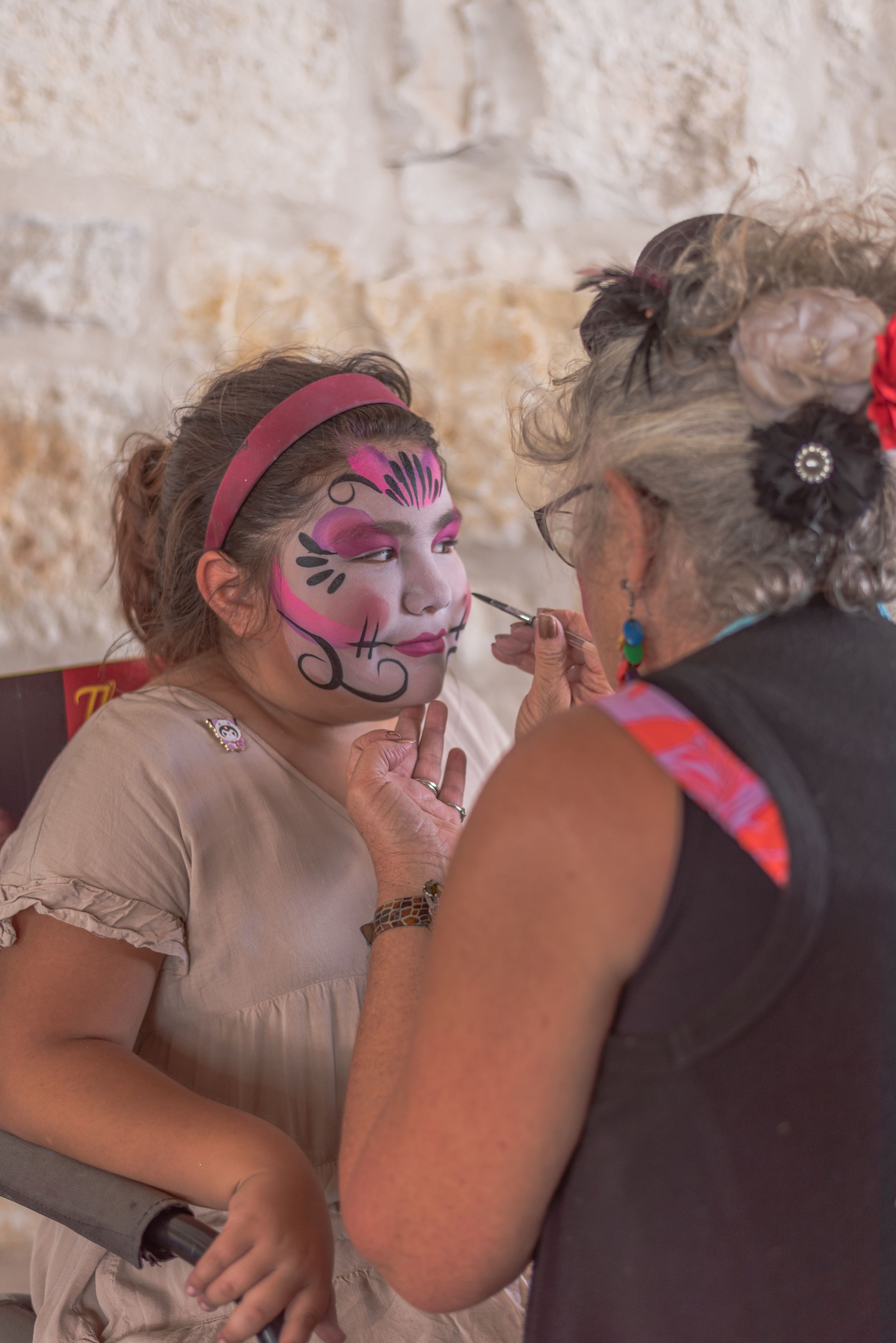A face painter applies colorful cat makeup on a young girl at a festive outdoor event.