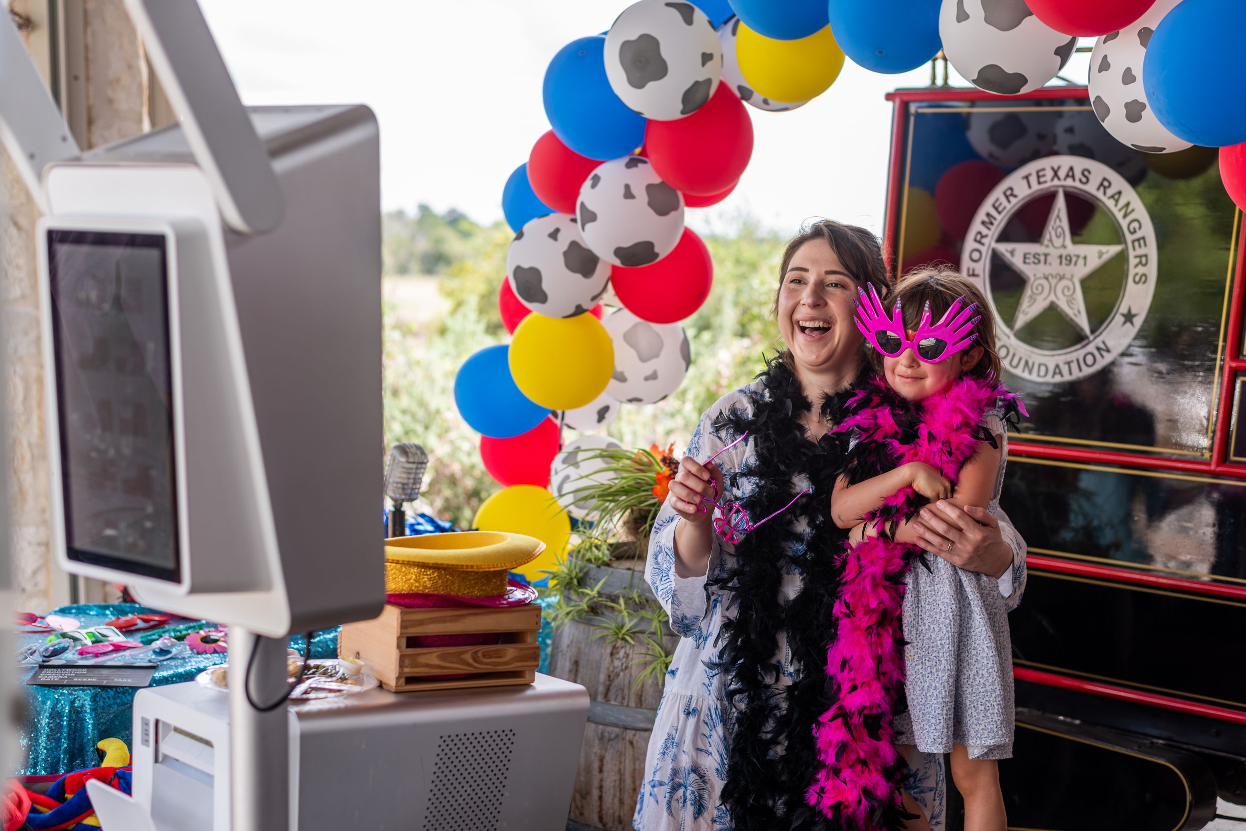 Woman and child laughing at a colorful photo booth with balloons and feather boas at a festive party.