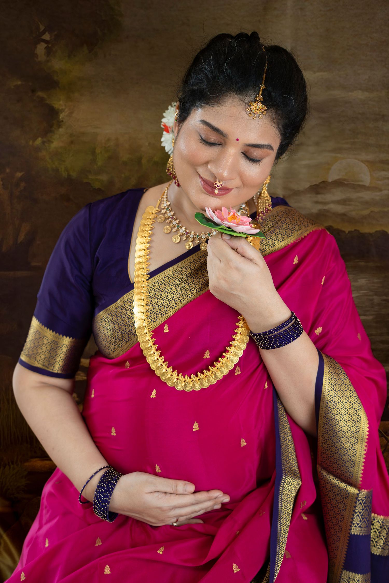Expecting mother in a pink Banarasi saree holding a lotus flower, eyes closed in a close-up maternity portrait
