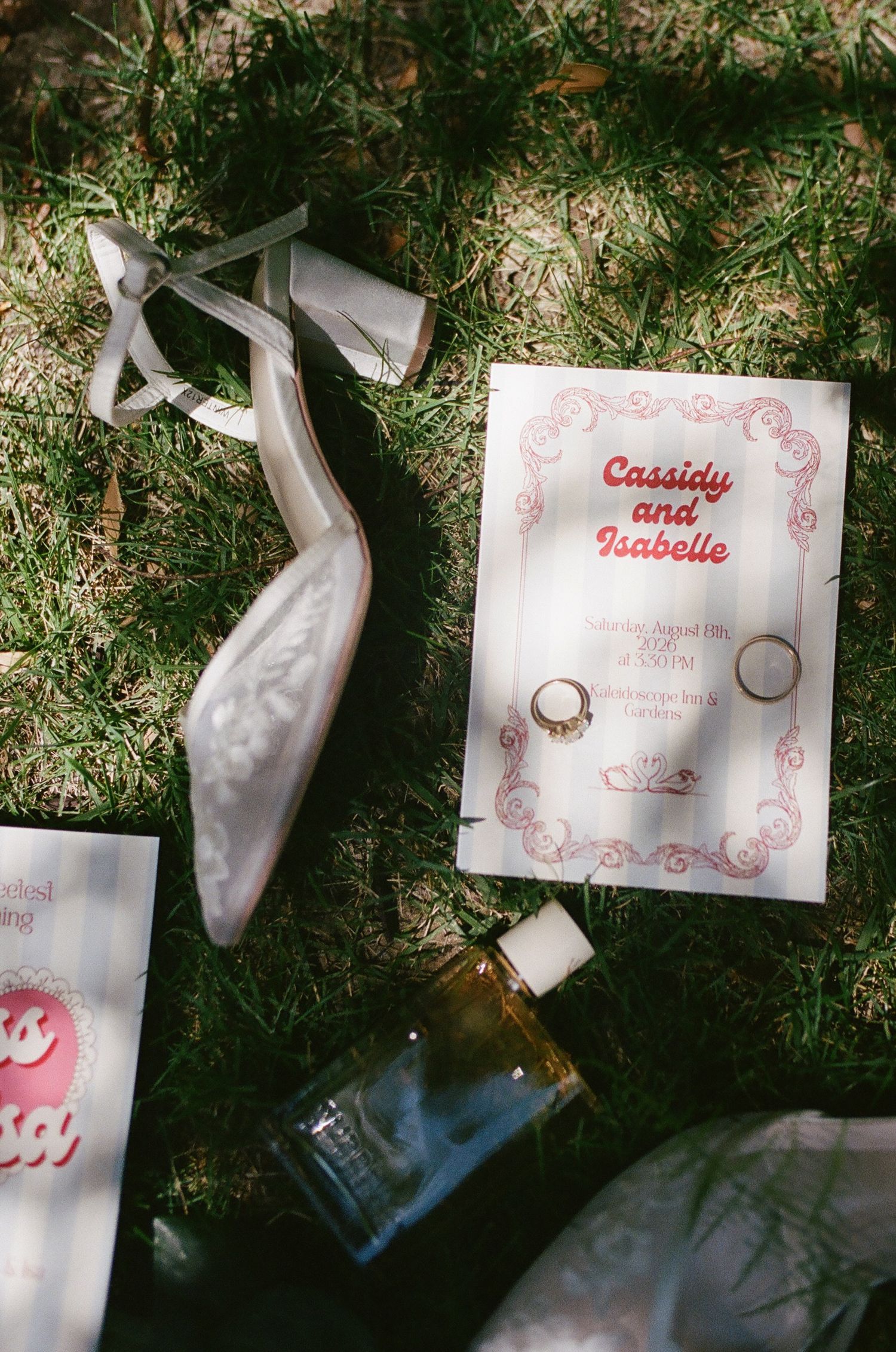 Wedding invitation for Cassidy and Isabelle with two gold rings, a white satin heel, and a perfume bottle resting on sunlit grass.