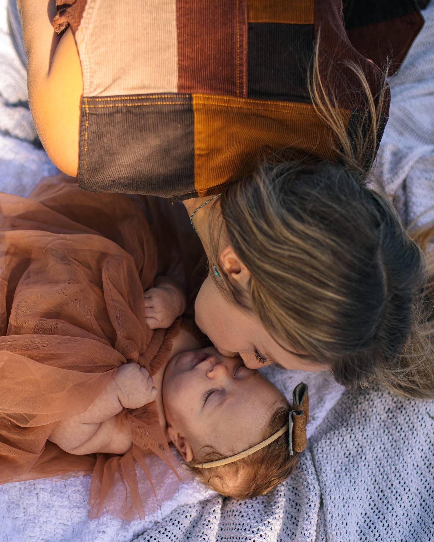 Mother in a patchwork top kisses the forehead of a sleeping baby in a rust tulle dress and headband on a soft white blanket.