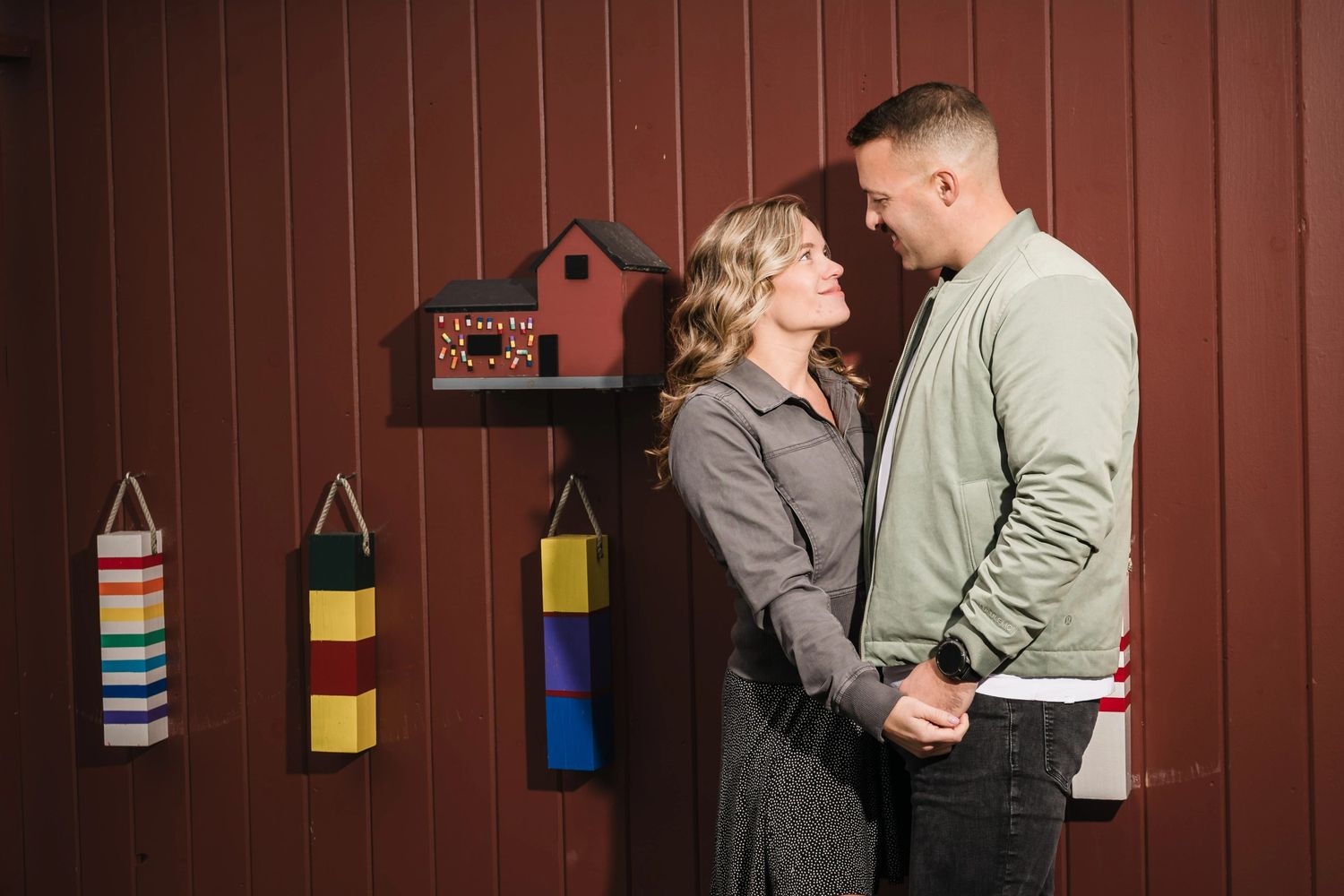 Couple gazing at each other while standing against a red corrugated metal wall with hanging decorative baskets. Engagement at Rockport MA
