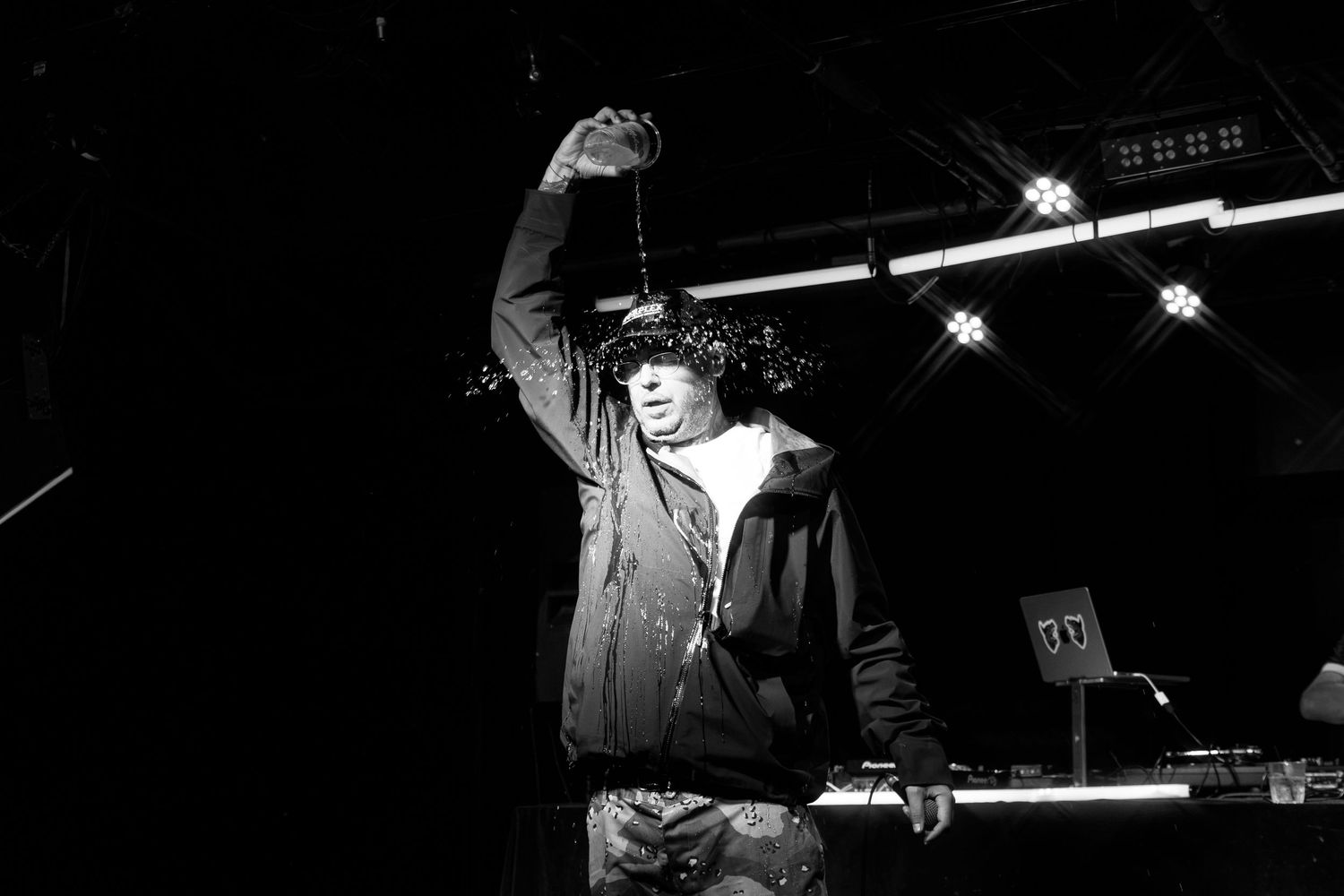 Black and white photo of a performer on stage holding an object above their head under bright stage lights.