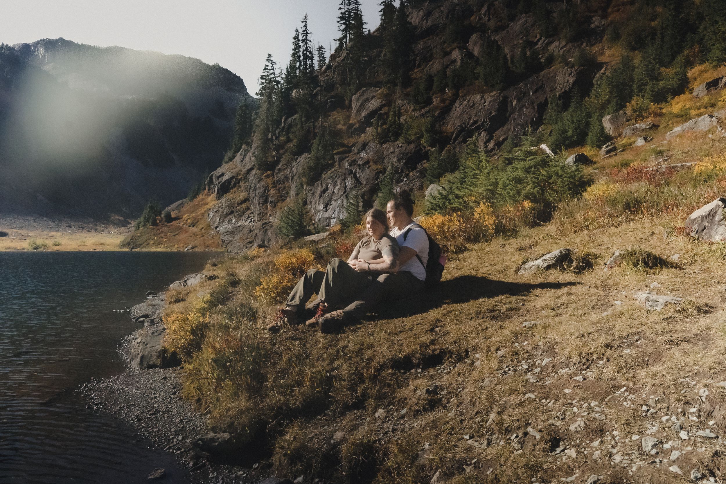 Couple sitting close together on a grassy hillside beside a calm alpine lake, surrounded by golden autumn foliage and rugged mountains.