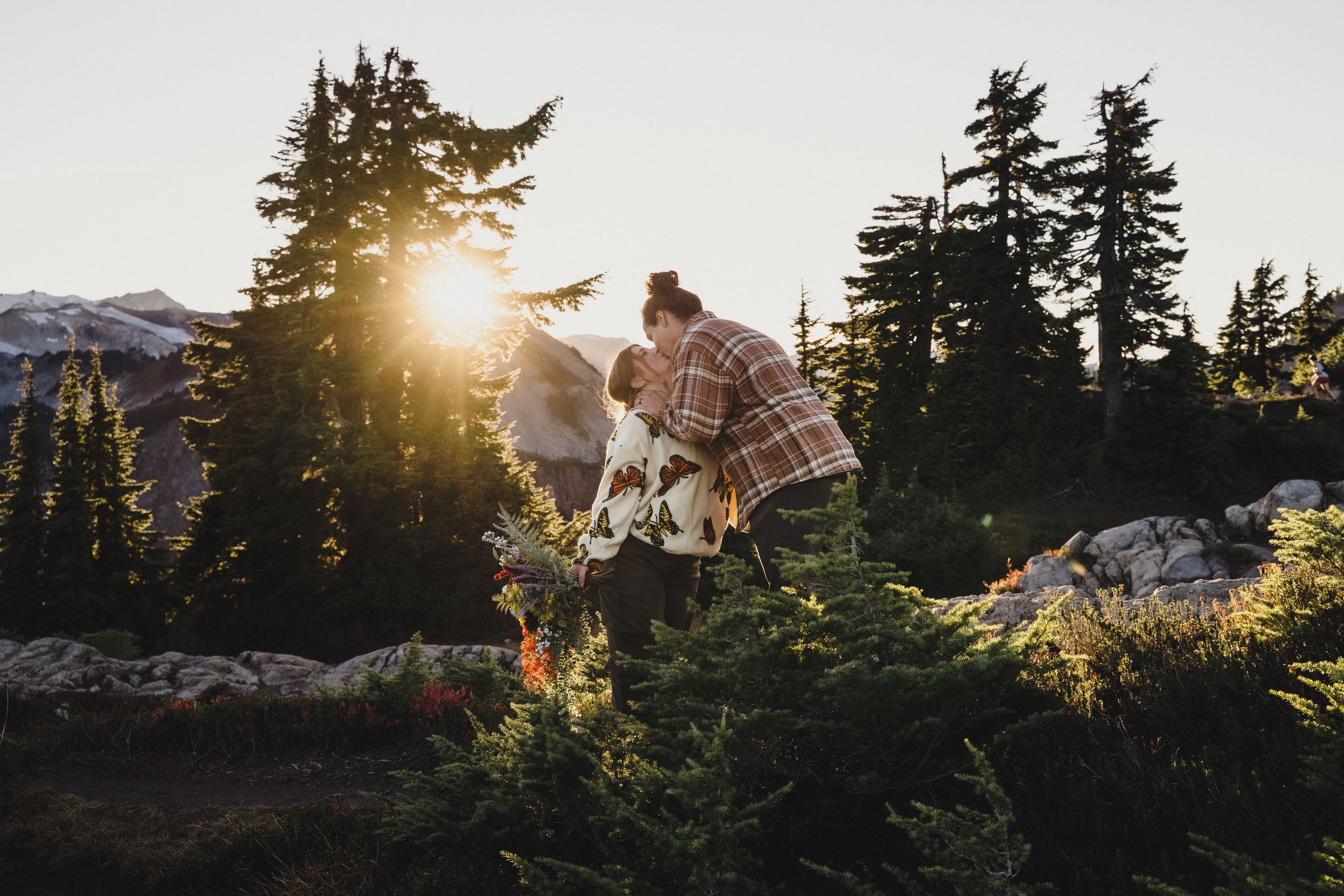 Couple embracing among evergreen trees and wildflowers on a mountain trail as golden sunset light glows over distant peaks.