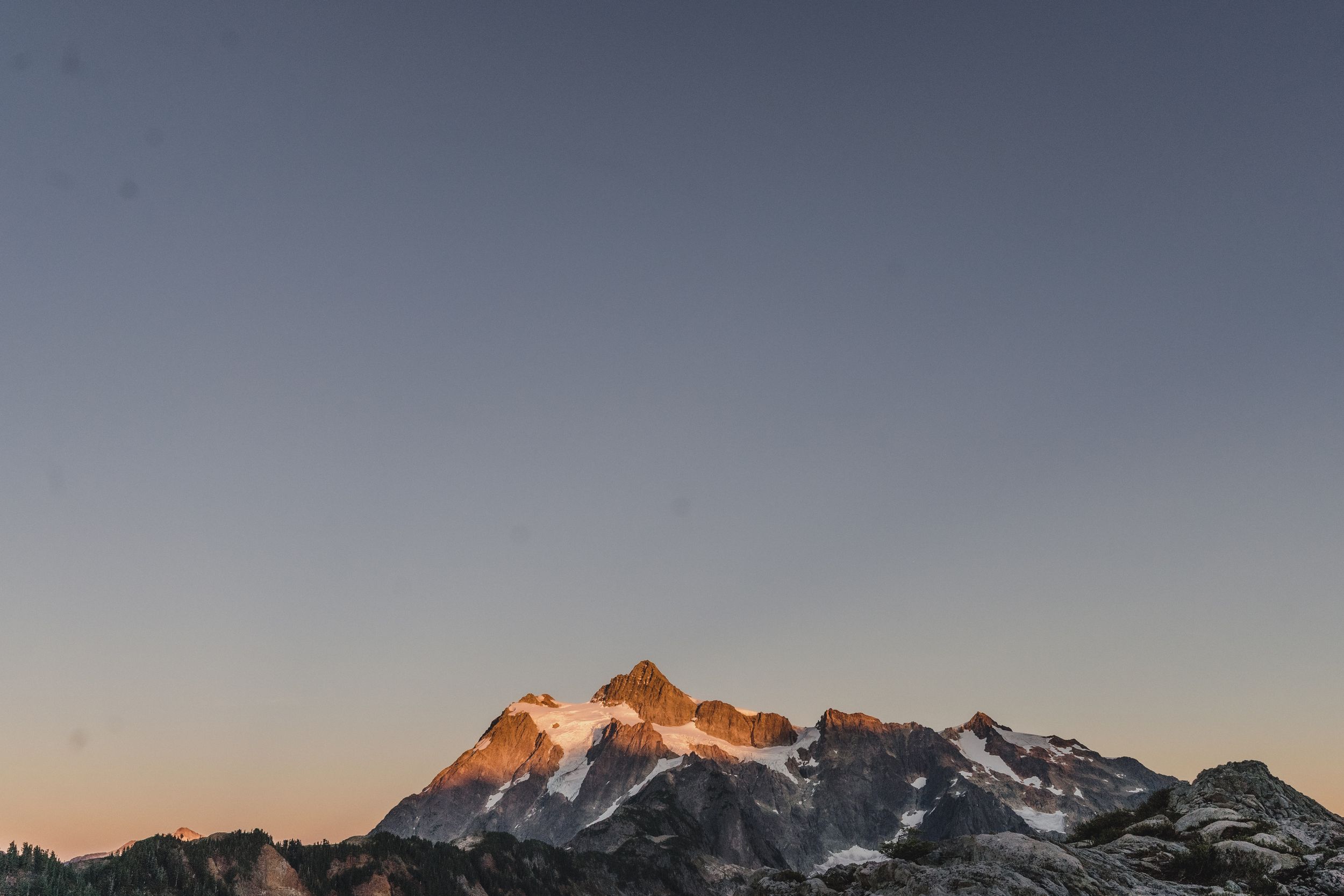 Snow-dusted mountain peak glowing orange in golden sunset light against a clear gradient sky above rugged rocky ridges.